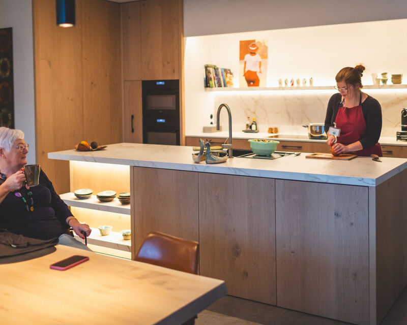 vrouwelijke medewerker snijdt groenten terwijl vrouwelijke zorgvrager koffie drinkt